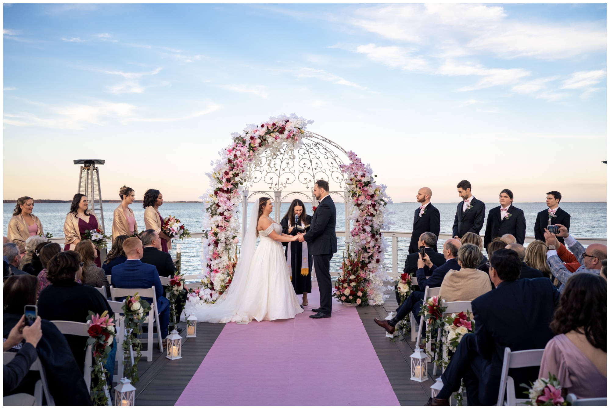 A couple stands at an outdoor altar decorated with pink and white flowers, exchanging vows at their celebrations at the bay wedding. Guests are seated on either side, overlooking a body of water under a blue sky, creating perfect wedding photos.