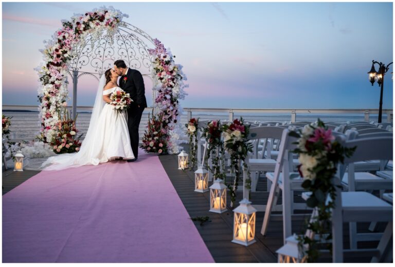 A bride and groom kiss under a floral arch at an outdoor ceremony by the sea, with rows of white chairs, lanterns, and flower arrangements lining a pink aisle at sunset—perfect for celebrations at the bay wedding photos.