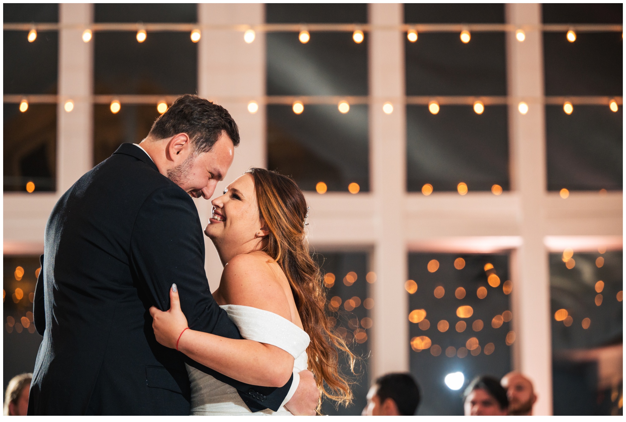 A bride and groom smile and embrace while dancing under string lights, with large windows and guests blurred in the background—perfect celebrations at the bay wedding photos.