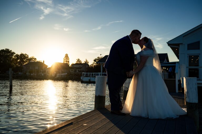 A bride and groom in wedding attire kiss on a dock at sunset, with water, houses, and trees in the background. The scene is warmly lit by the setting sun as an Anchor Inn wedding photographer captures their romantic silhouette.