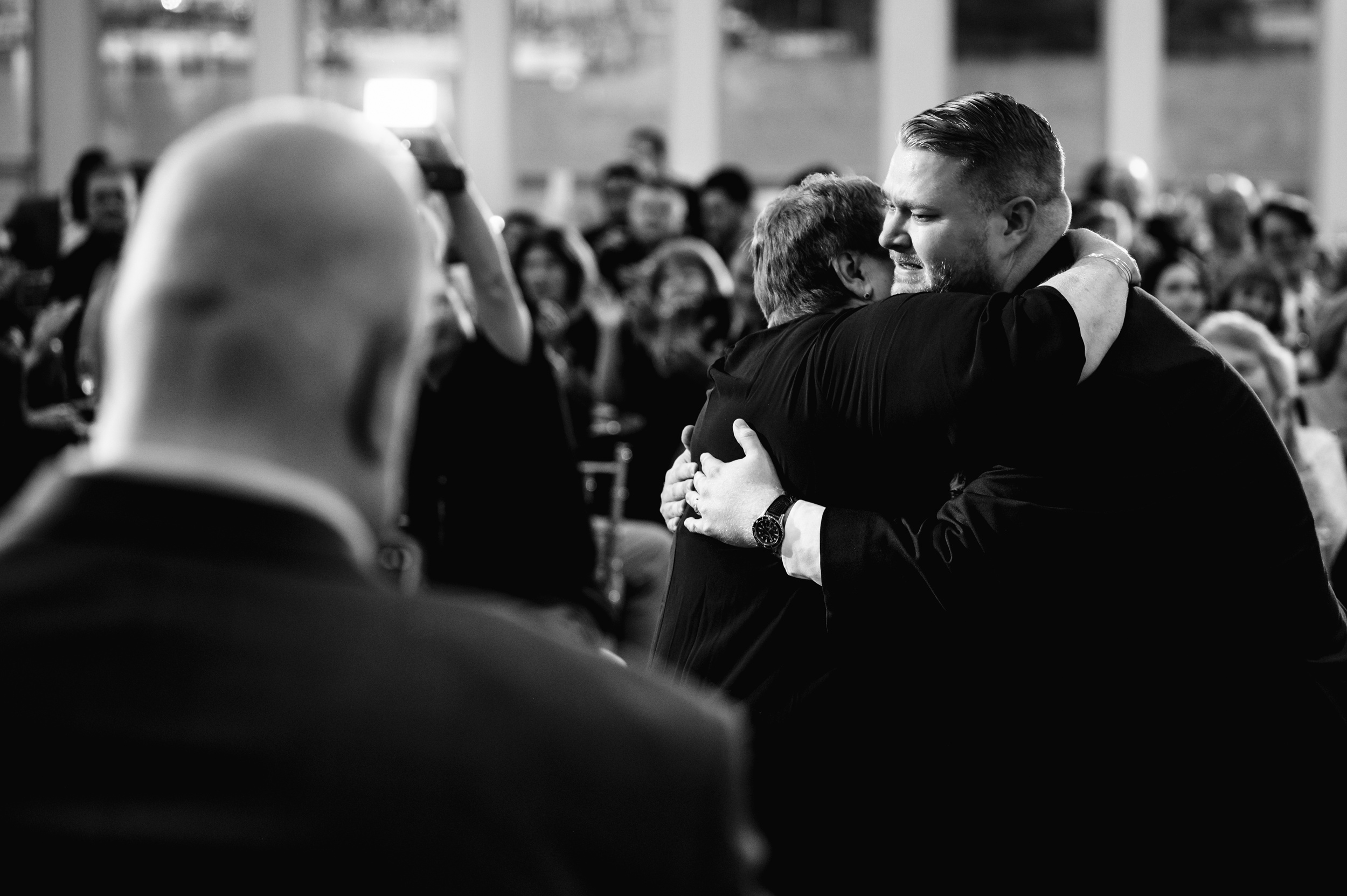 Mother and son hug emotionally in front of an audience at an Anchor Inn wedding, with one man's back to the camera and the crowd blurred in the background. A bald man is in the foreground, out of focus. The setting appears formal, possibly a ceremony.