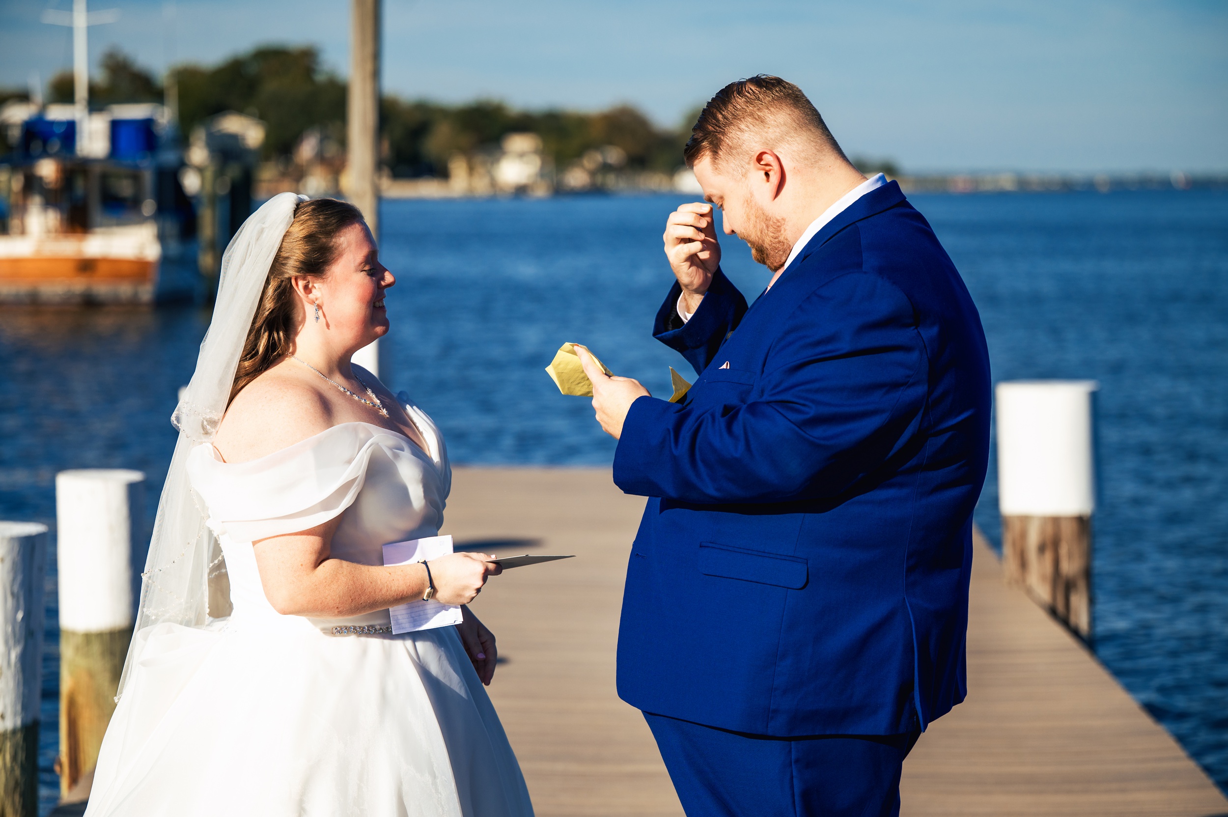 A bride and groom stand on a dock by the water. The groom, dressed in a blue suit, wipes his eyes with a tissue while reading a note. Captured by their Maryland wedding photographer, the bride smiles warmly, holding her vows in her hands.