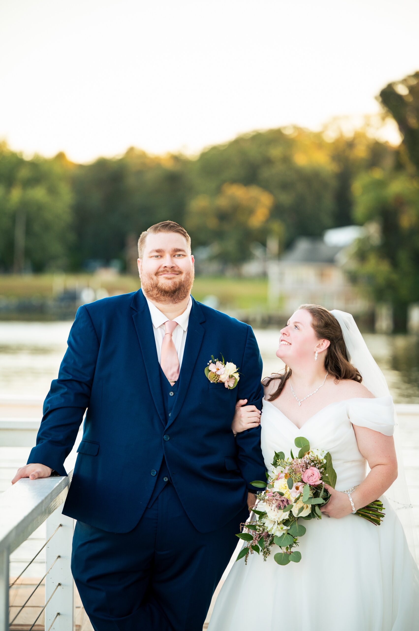 A bride in a white dress and veil holds a bouquet, smiling up at her groom in a navy suit with a pink tie. Captured by an Anchor Inn wedding photographer, they stand outdoors by the water, trees and houses in the background.